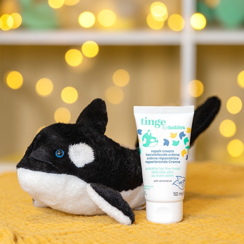 Weiße Tube "tinge for babies Repair Creme" neben einem Orca-Plüschtier. Produktbezeichnungen und 50 ml-Angabe sichtbar.