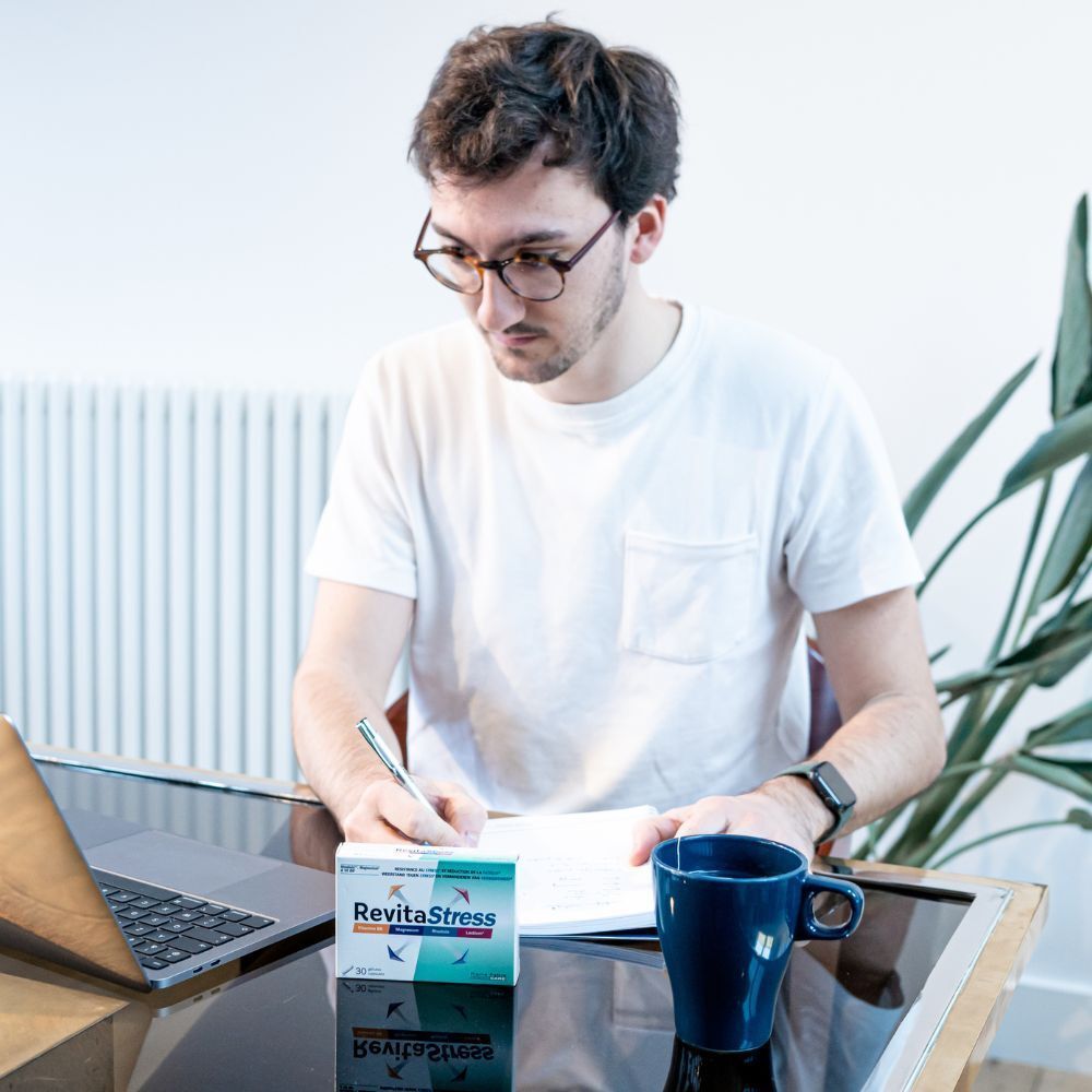 Homme au bureau avec ordinateur portable, carnet et boîte "RevitaStress". Il boit du thé.