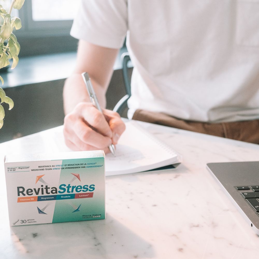 Personne écrit dans un carnet, à côté d'une boîte "RevitaStress". Ordinateur portable en arrière-plan.