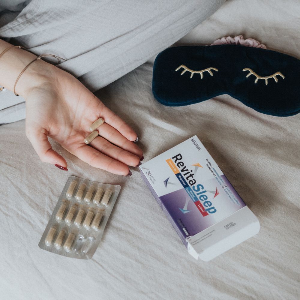 Hand met capsules en een verpakking RevitaSleep. Daarnaast een blisterverpakking met capsules en een slaapmasker op beddengoed.