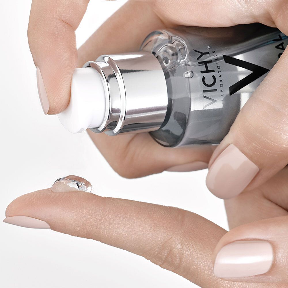 Main pressant le produit du flacon Vichy Mineral 89. Liquide clair sur le doigt. Pompe argentée, ongles rose clair.