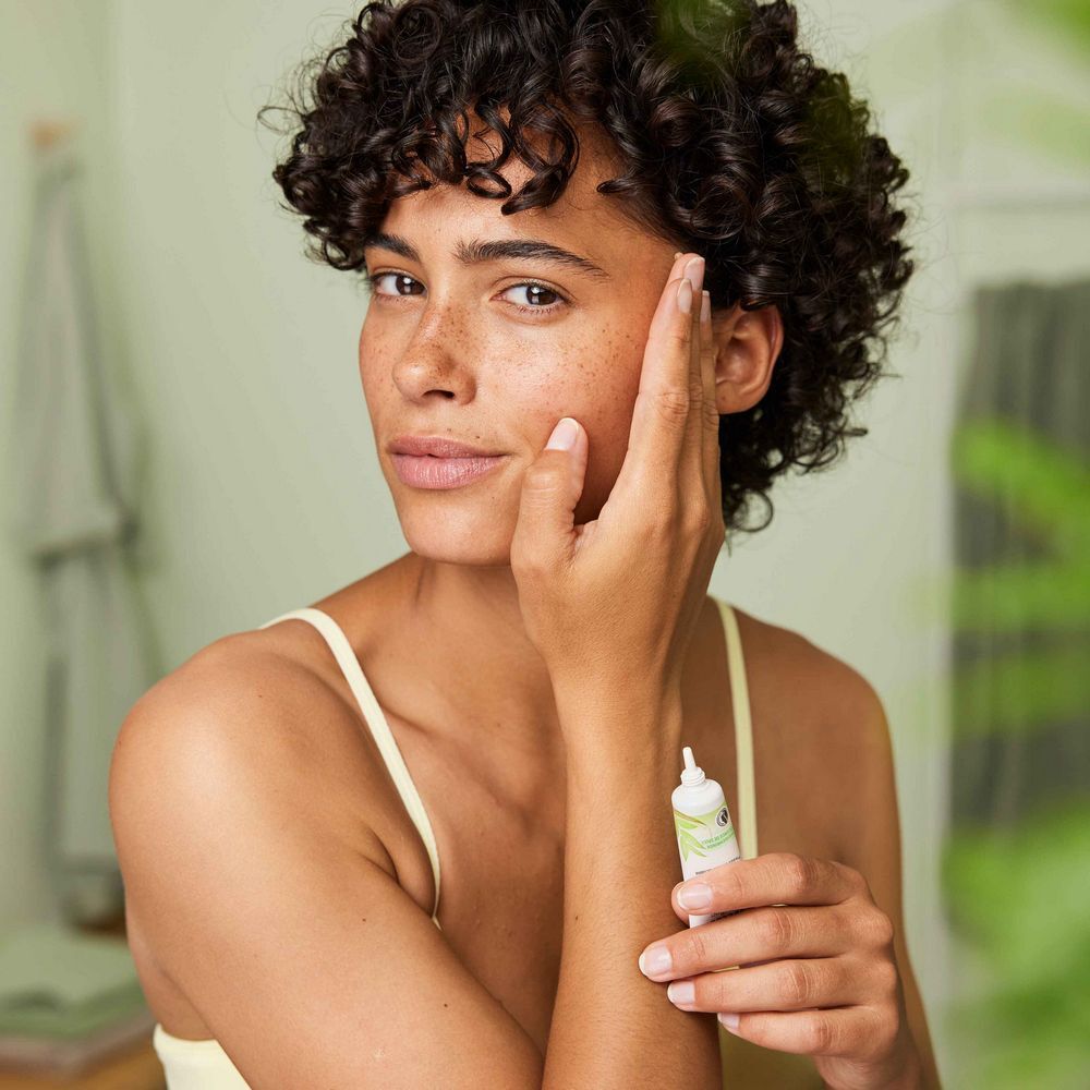 Femme tenant un petit tube devant son visage. Peau claire, cheveux bouclés. Porte un haut jaune. Tient un tube avec des accents verts.