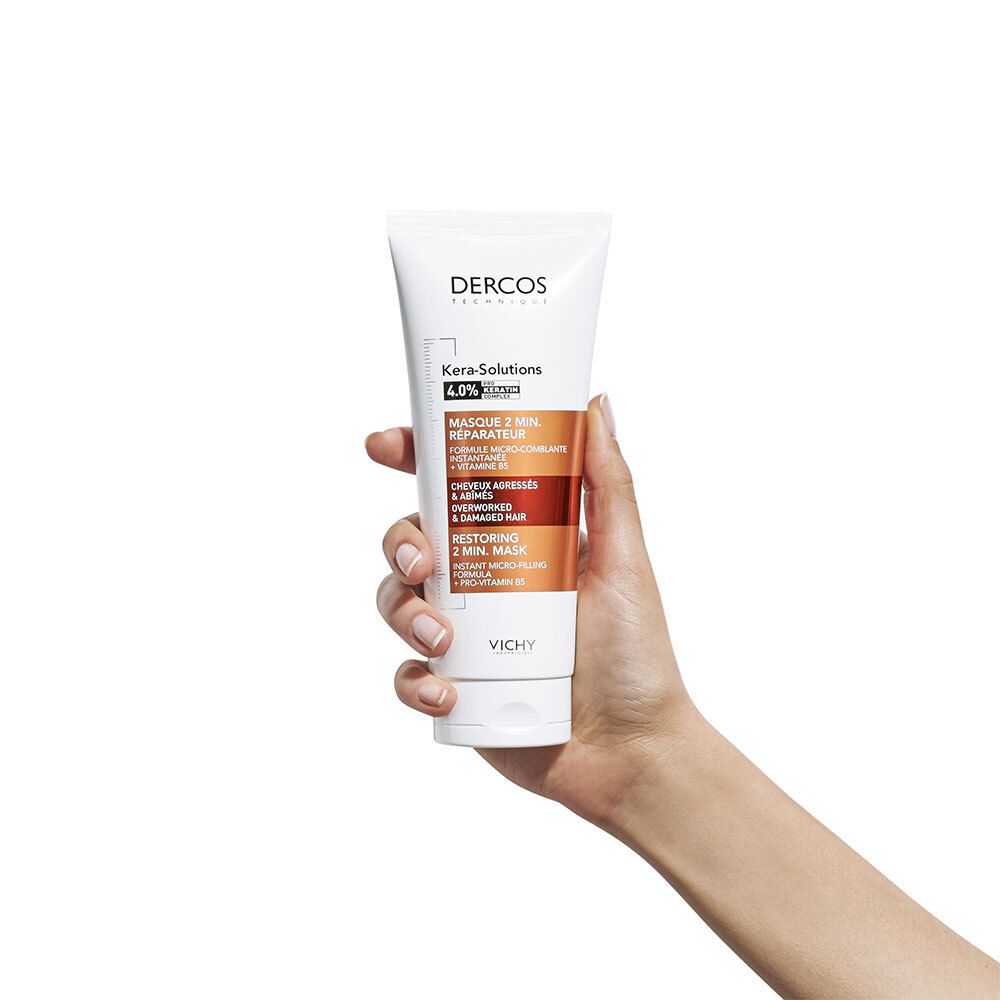 Main tenant un tube blanc. Inscriptions: Dercos, Kera-Solutions, Masque 2 min. réparateur. Accents marron et rouge.