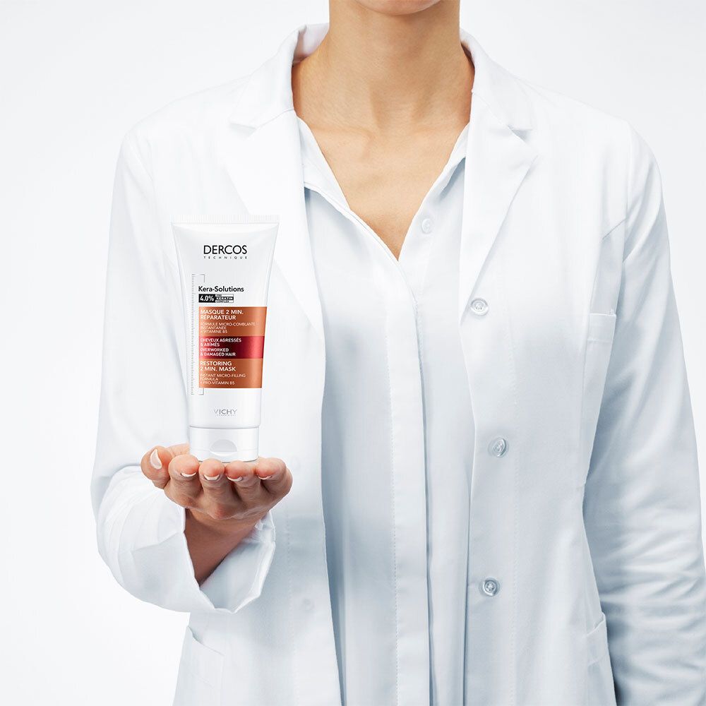 Personne en blouse blanche tenant un tube. Inscriptions: Dercos, Kera-Solutions, Masque 2 min. réparateur. Accents marron et rouge.
