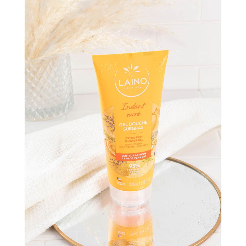 Tube jaune sur un miroir, entouré de décorations. Inscriptions : LAINO, Instant sucré, Gel Douche Surgras, 95% ingrédients d'origine naturelle.