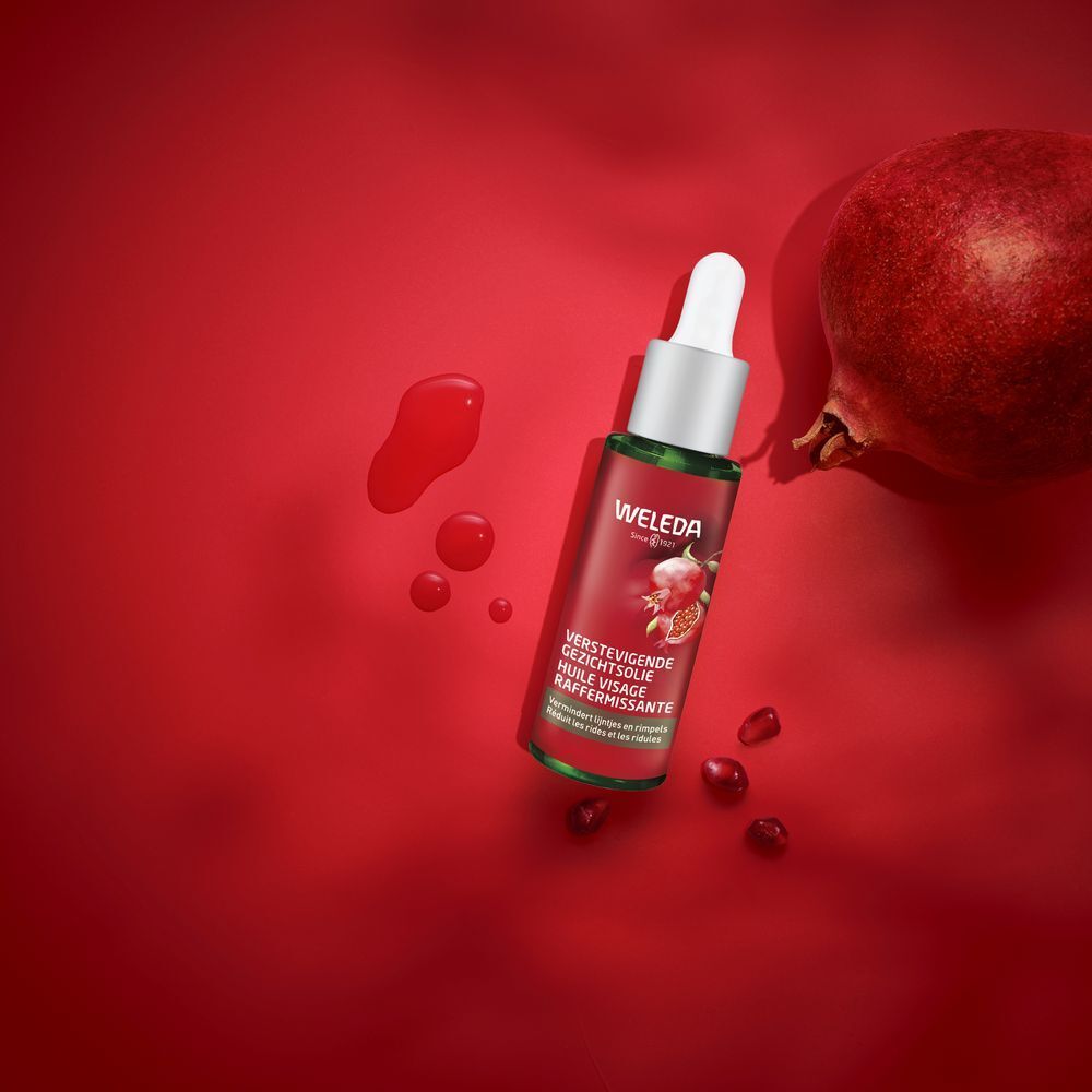 Flacon d'huile pour le visage avec pipette et grenade sur fond rouge. Logo WELEDA et nom du produit visibles.