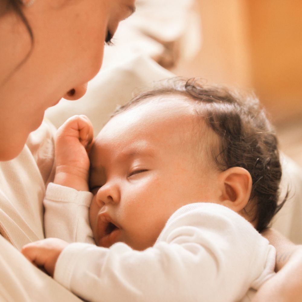 Nahaufnahme eines schlafenden Babys im Arm einer Person. Baby in weißem Strampler. Person nicht vollständig sichtbar.