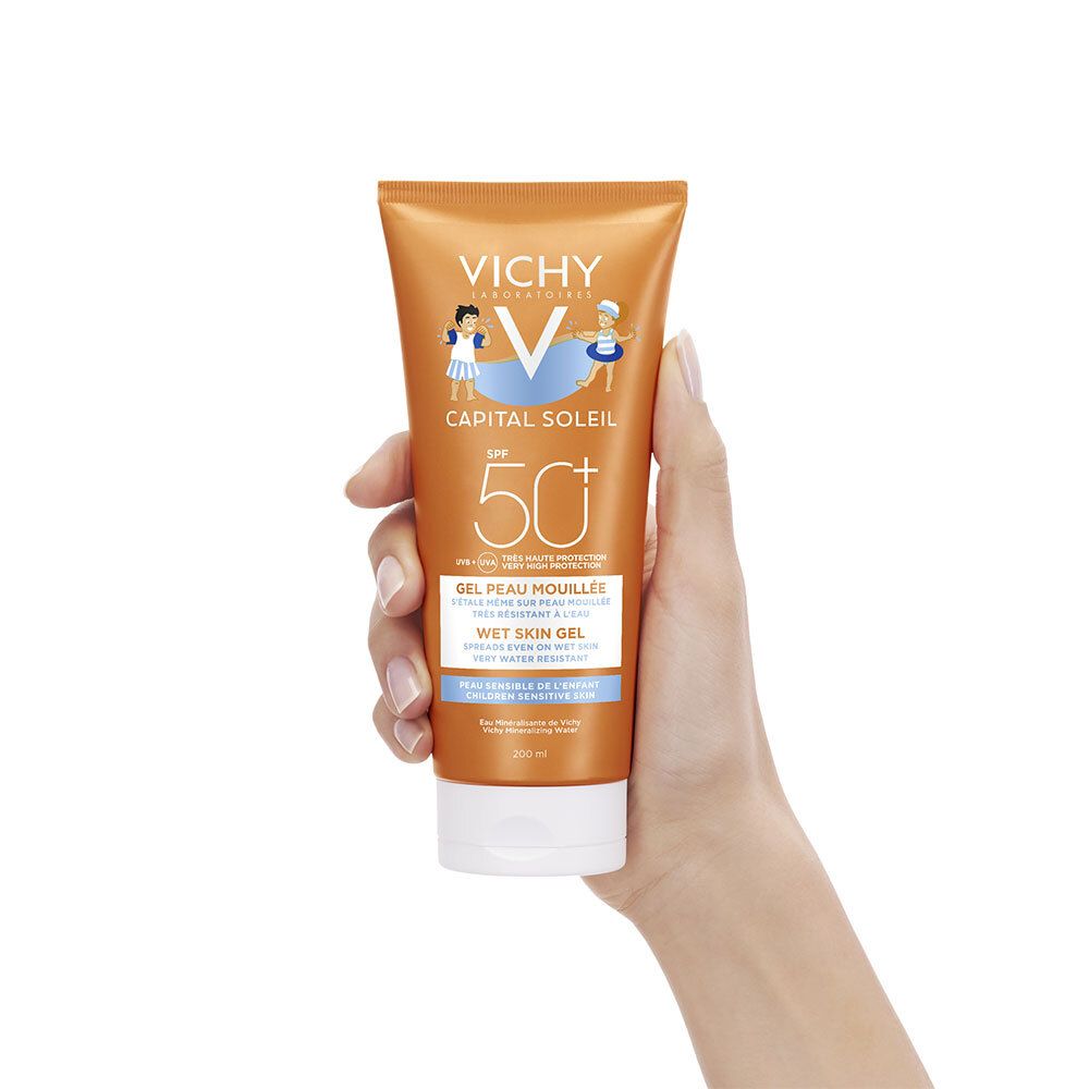 Main tenant un tube de crème solaire. Marque Vichy, SPF 50+. Texture gel, pour peau mouillée. Présentation du produit.