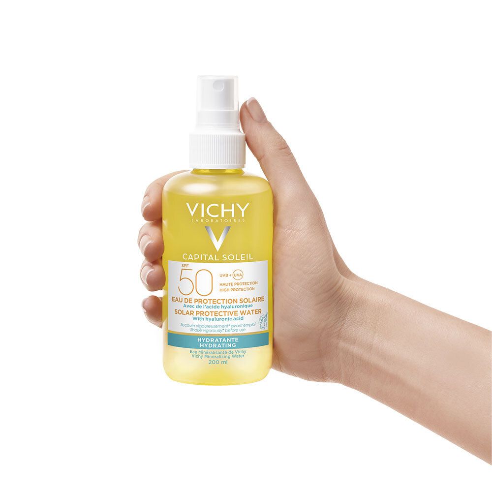 Hand houdt gele fles met verstuiver. Vichy Capital Soleil SPF50, tekst.