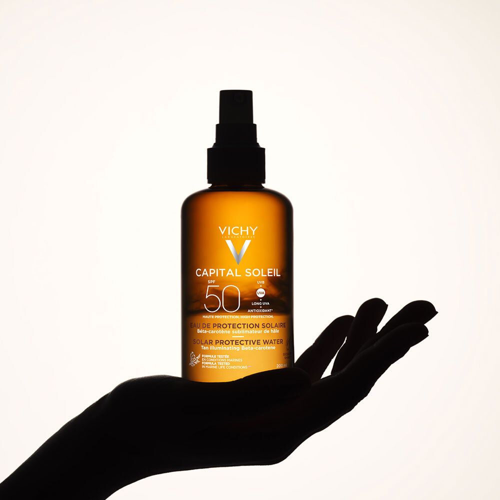 Silhouette d'une main tenant un flacon Vichy Capital Soleil Eau de protection solaire SPF50.
