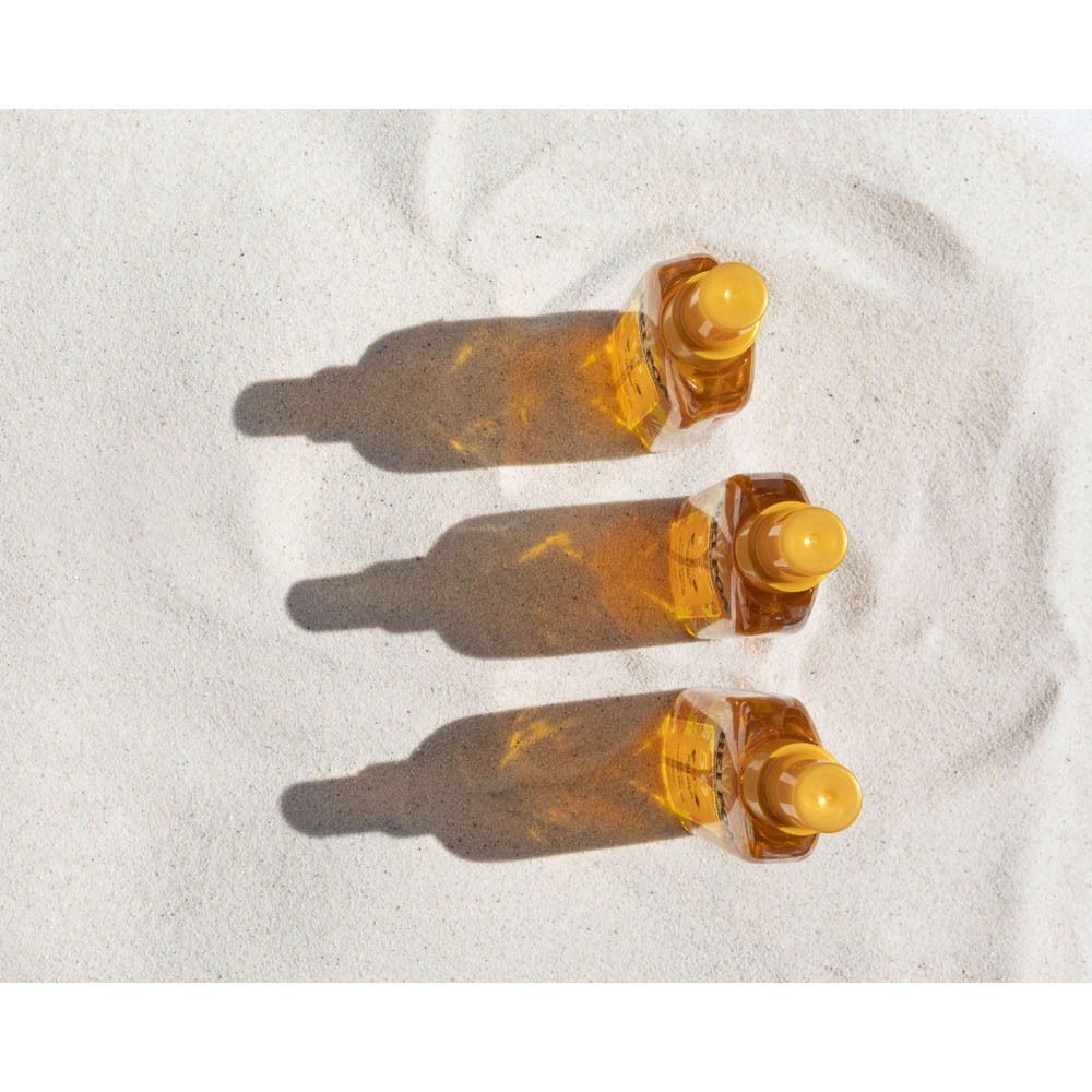 Trois flacons spray oranges avec bouchons jaunes sur du sable. Ombres visibles.