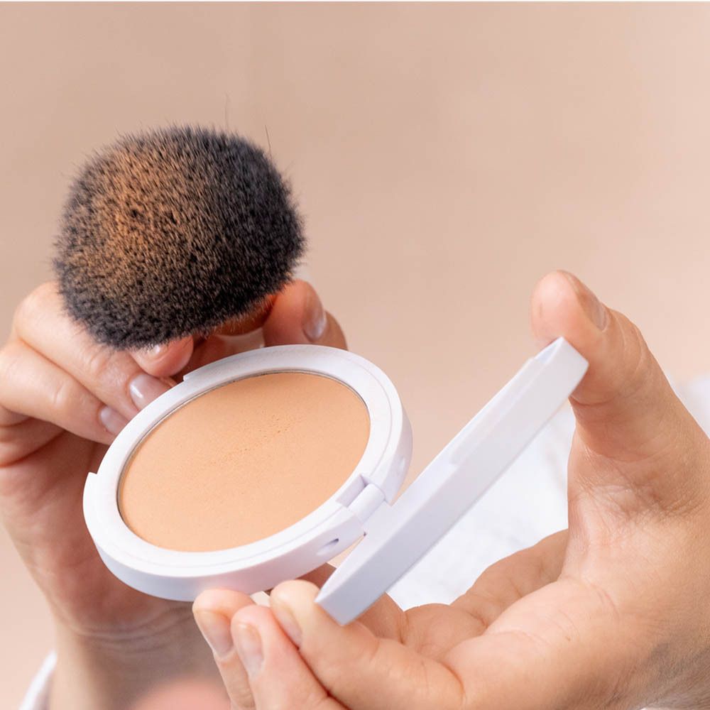 Persoon houdt poederdoos en kwast vast. Beige poeder. Kwast met zwart haar.
