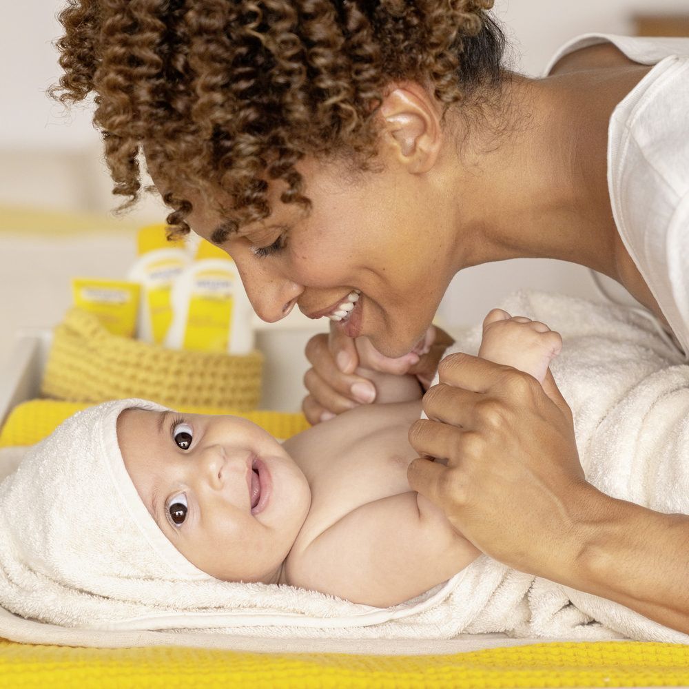 Een moeder en een baby. De baby ligt op een gele ondergrond, gewikkeld in een handdoek. De moeder lacht.