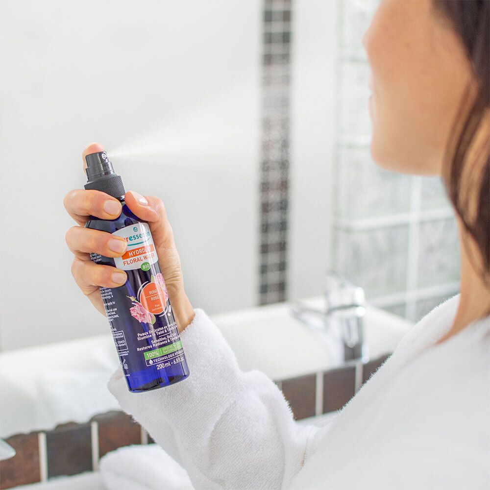 Persoon spuit met een blauwe fles. Opschrift: Puressentiel, Hydrolat Floral Water Bio. Spiegel op de achtergrond.