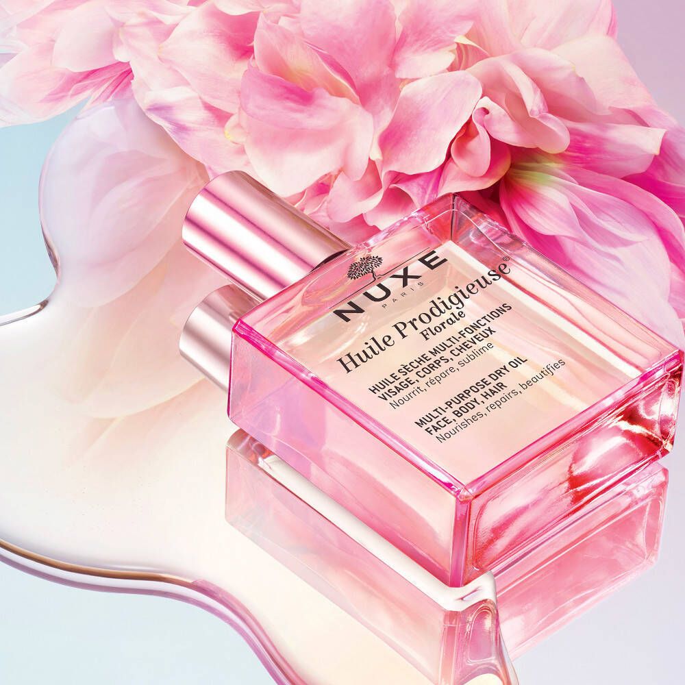 Glazen fles met roze dop, omringd door roze bloemen. Opschrift: NUXE Huile Prodigieuse Florale. Product op glanzend oppervlak.
