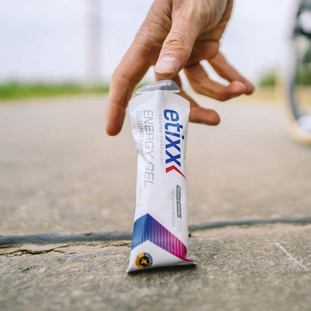 Main tenant une sachet Etixx Energy Gel. Inscription : Energy Gel, boisson isotonique. Déchirure grise.