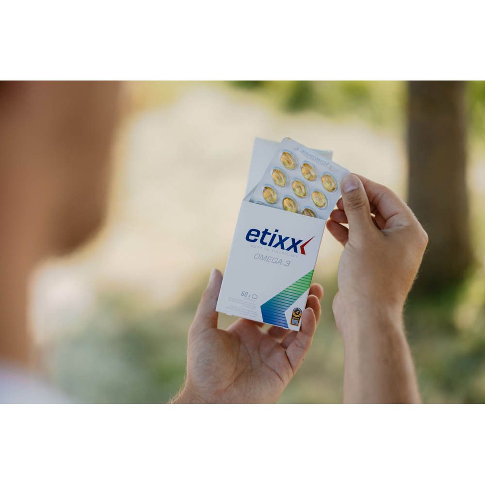 Hände halten eine geöffnete Packung "etixx OMEGA 3" mit Kapseln. Hintergrund: verschwommene Natur. Verpackung mit Logo.