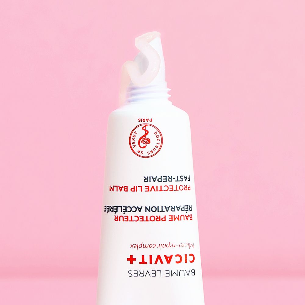 Gros plan d'un tube de baume à lèvres ouvert. Fond rose. Nom du produit et logo visibles.