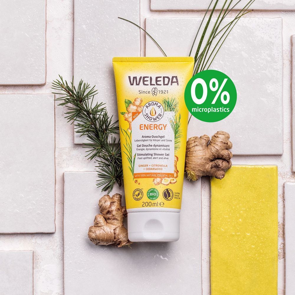 Gele tube "WELEDA ENERGY" op witte achtergrond. Gember en groene takken decoreren de scène. 0% microplastics.
