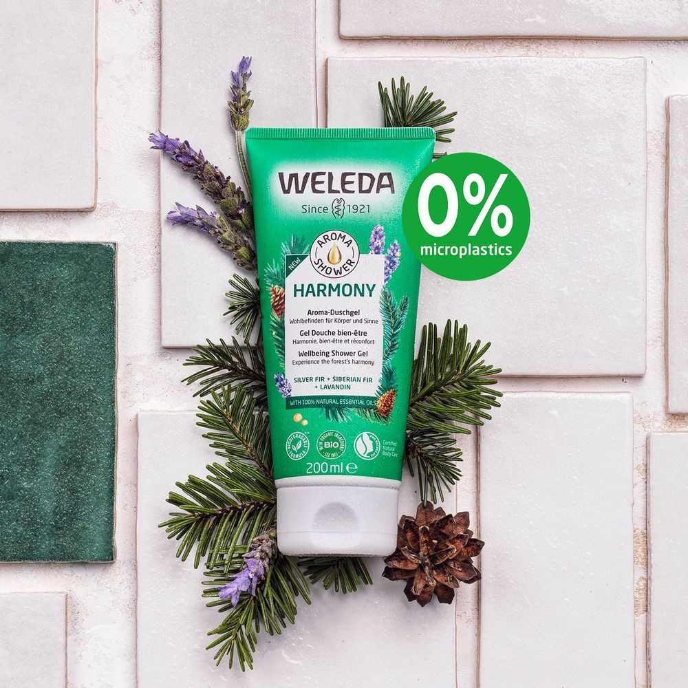 Groene tube met witte dop, omringd door lavendel, dennentakken en dennenappels. Opschrift: WELEDA, Aroma Shower Harmony. Met 0% microplastics label.