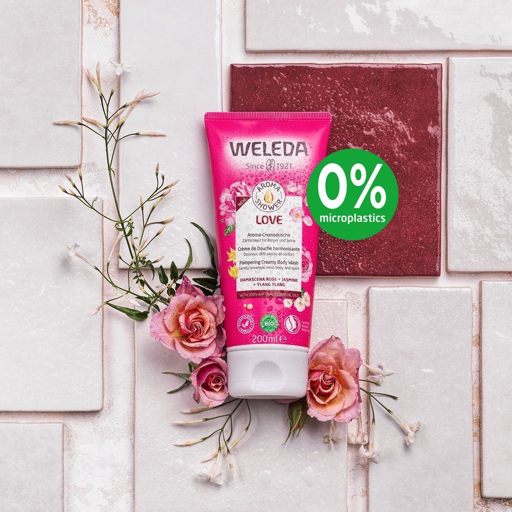 Roze tube "WELEDA" en "LOVE" op tegels. Rozen en bloemen omringen de tube. 0% microplastics label.