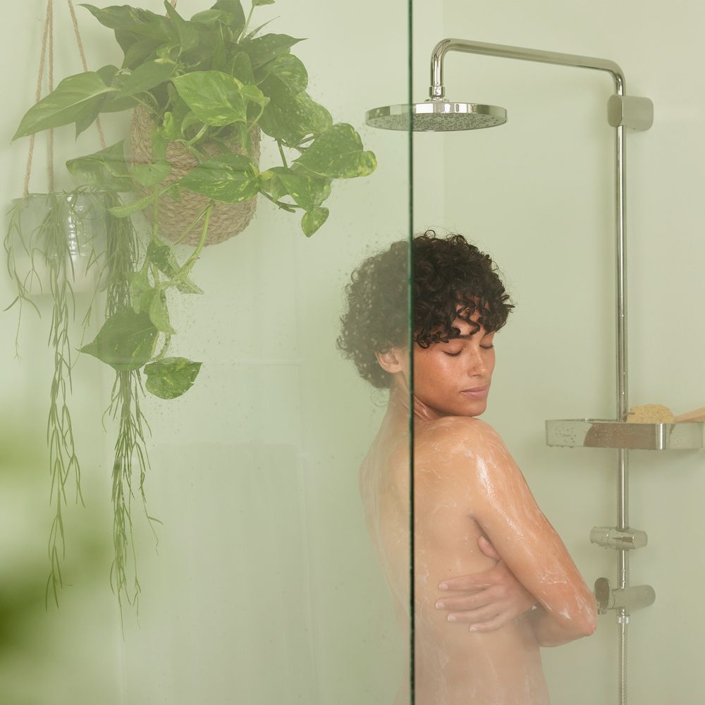 Persoon onder de douche, ogen dicht. Douchekop en planten op de achtergrond. Glazen wand.