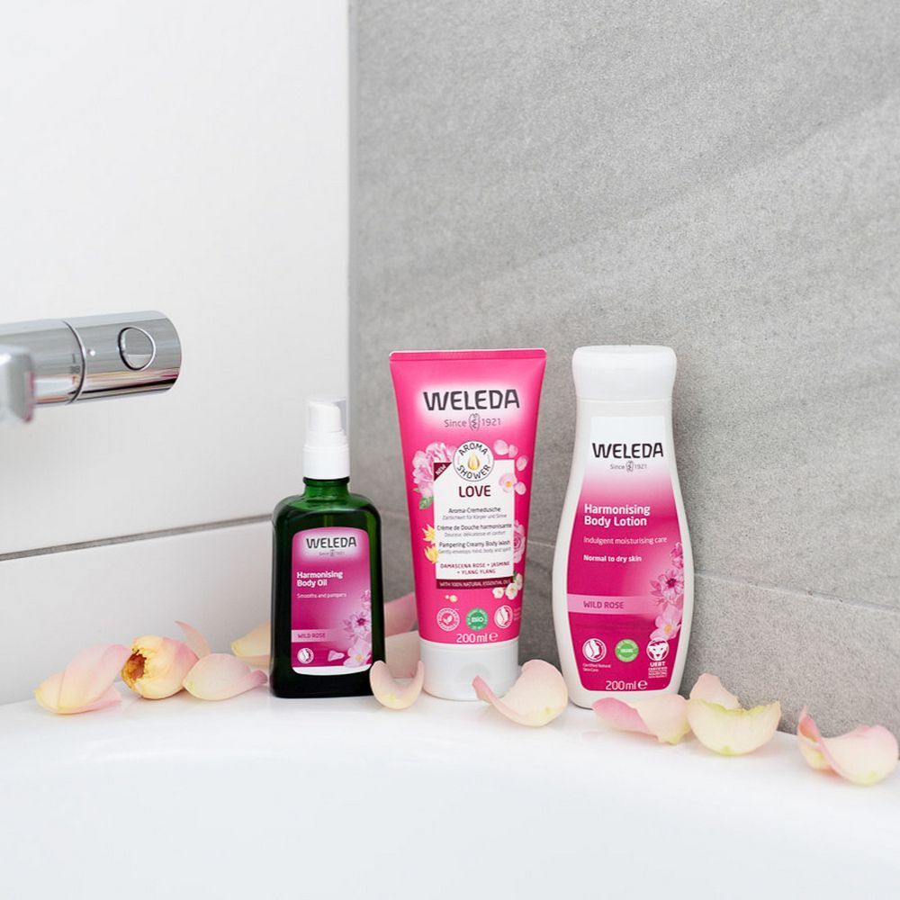 Drie Weleda-producten op een oppervlak. Roze tube, groene fles, witte fles. Rozenblaadjes.