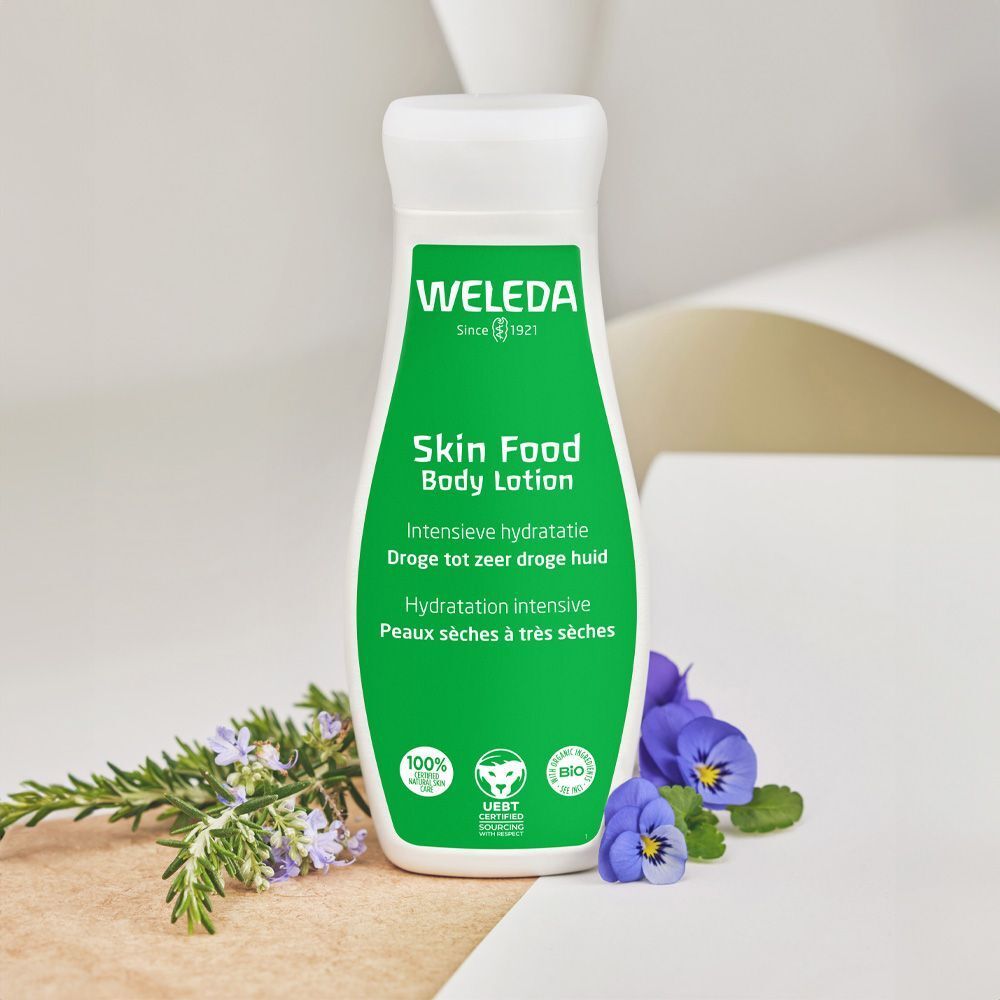 Groene fles met witte dop. Opschrift: Weleda Skin Food Body Lotion. Bloemen en tak ernaast.