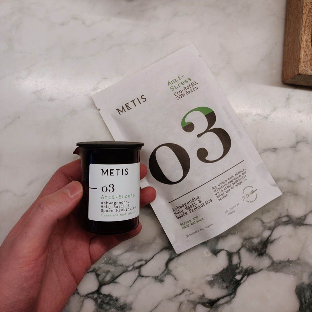 Hand houdt een zwarte pot en een witte zak vast. Pot met METIS-logo en "O3"-opdruk. Zak: Anti-Stress, Eco-Refill, 20% Extra.