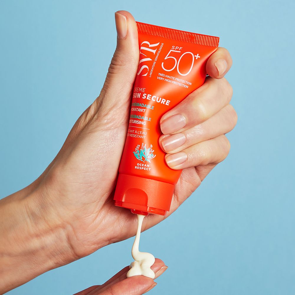 Main tenant un tube orange SVR Sun Secure Crème SPF50+. Crème appliquée sur le doigt.