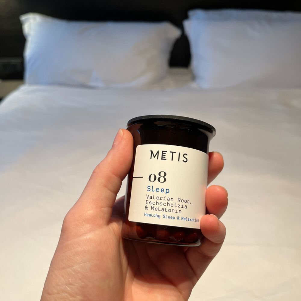 Hand houdt een bruine glazen fles met zwarte deksel. Label met tekst: METIS 08 Sleep. Capsules zichtbaar.