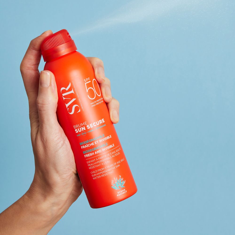 Main tenant un flacon spray orange. Brouillard de pulvérisation. Inscriptions: SVR, SPF 50, SUN SECURE.