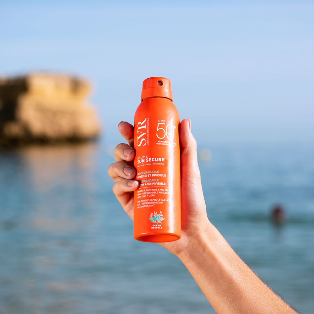 Main tenant un flacon spray orange. Arrière-plan : mer. Inscriptions: SVR, SPF 50, SUN SECURE.