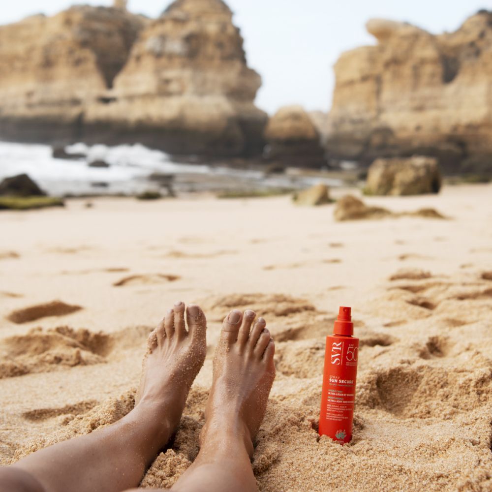 Pieds sur la plage à côté d'un flacon spray orange. Inscriptions : SVR, SUN SECURE Spray SPF50+.