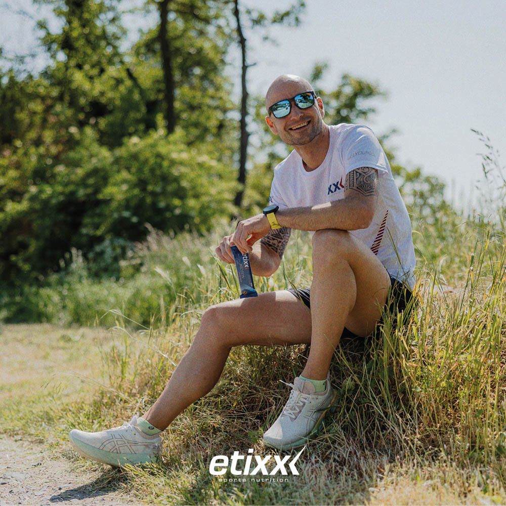 Man met zonnebril houdt eiwitreep vast. Opschrift: etixx. Achtergrond: bomen en gras.