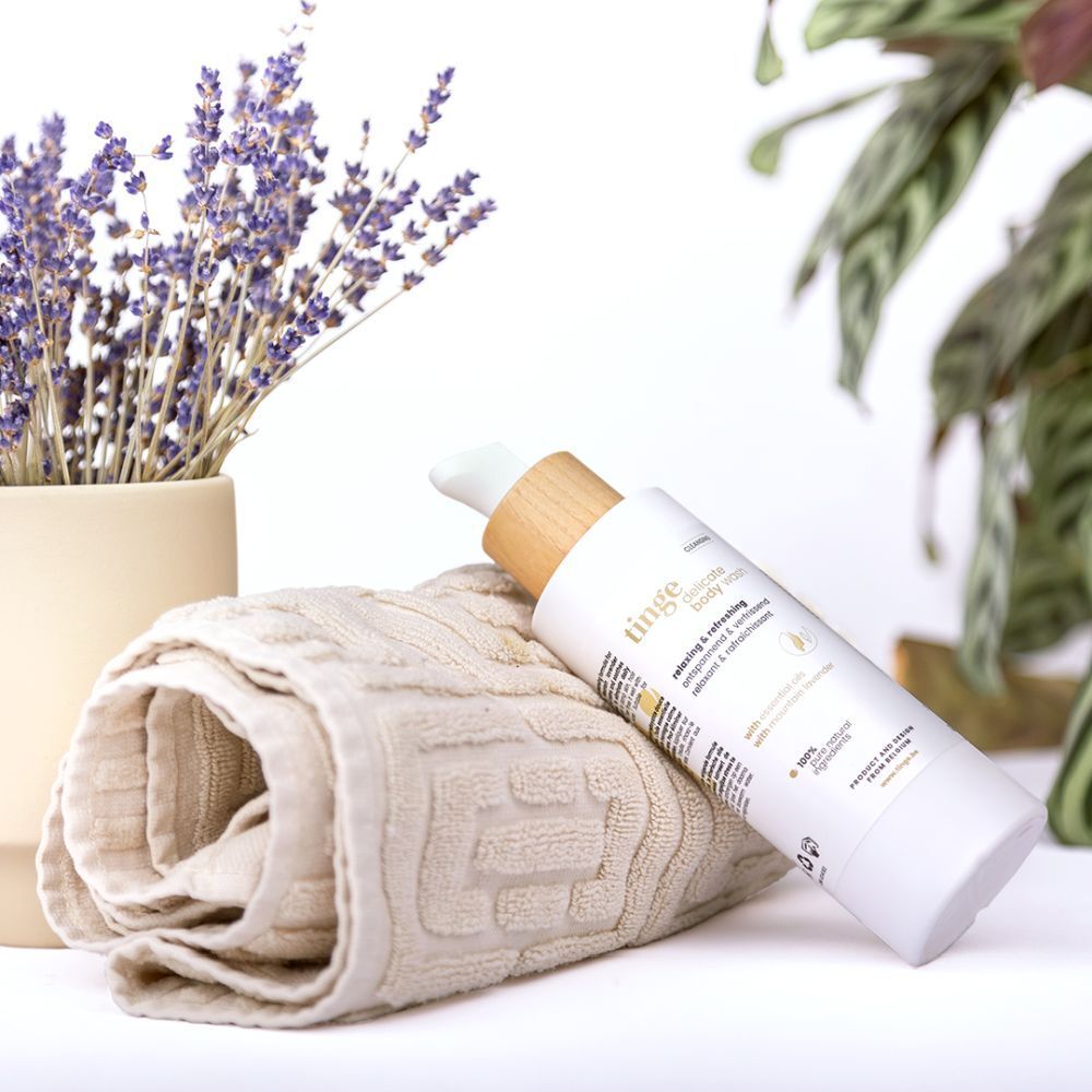 Weiße Flasche mit Pumpe und Holzdeckel neben Handtuch und Lavendel. Aufschrift: tinge delicate body wash.