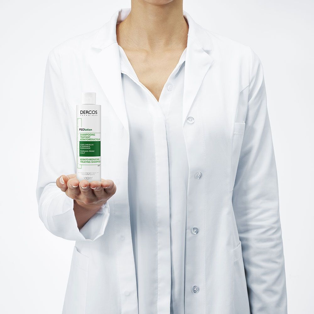Persoon houdt een witte fles vast. Tekst: Dercos, PSolution, Shampooing Traitant Kératoréducteur. Witte jas.