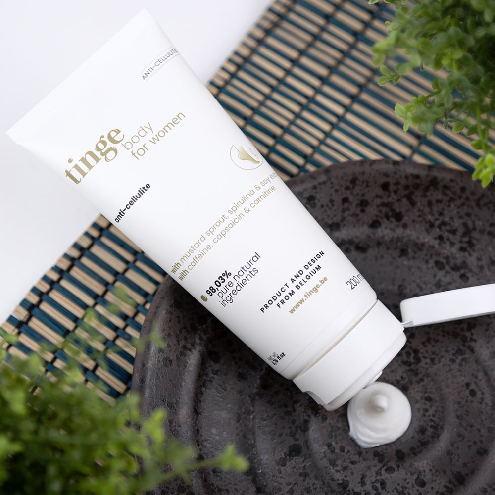 Weiße Tube, Produktbeschriftung: tinge body for women, anti-cellulite, 98,03% natürliche Inhaltsstoffe. Creme auf Teller. Pflanze im Hintergrund.