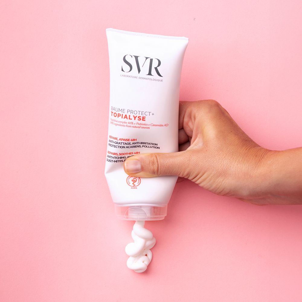 Hand houdt een tube vast, waar crème uit komt. Roze achtergrond. Productnaam zichtbaar.