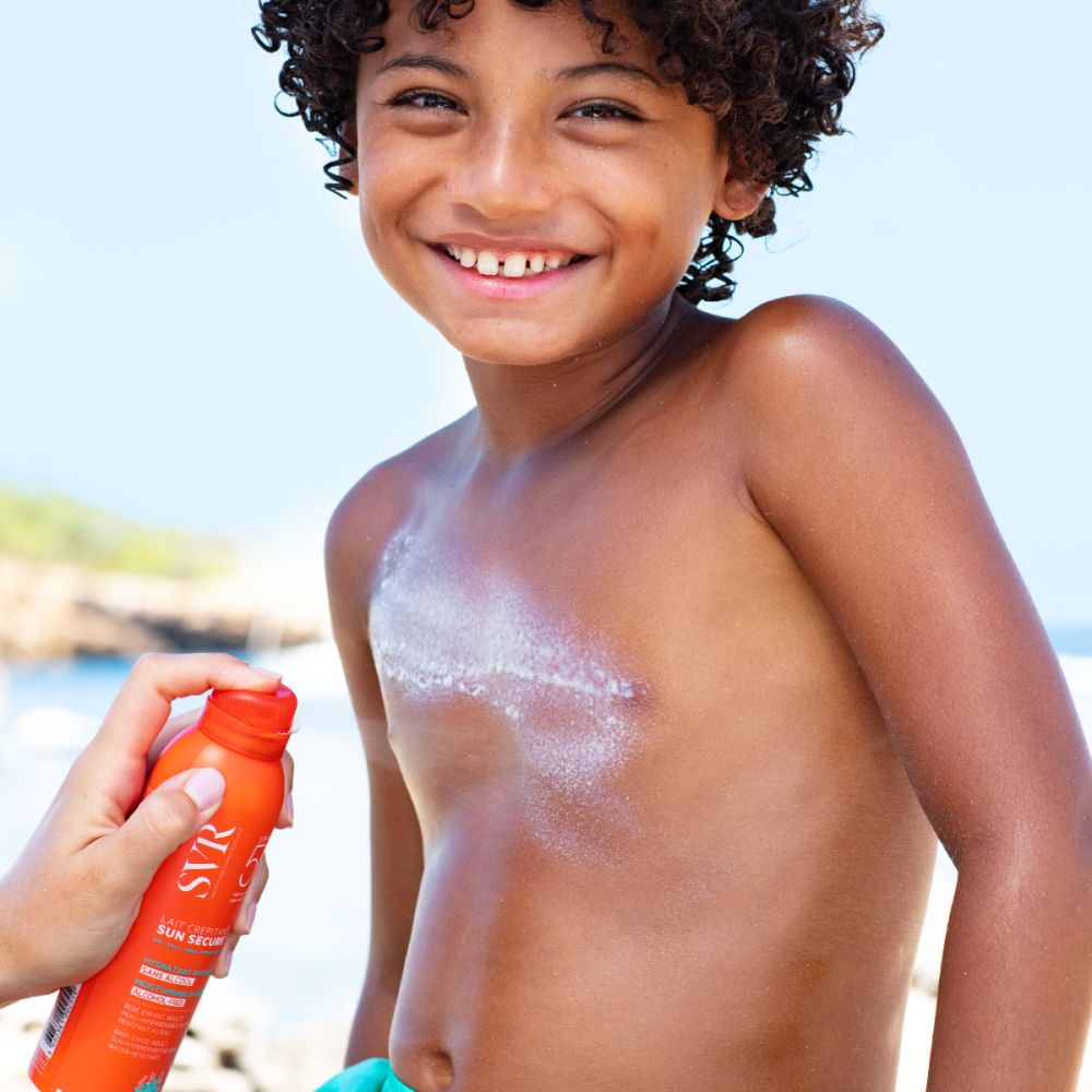 Enfant se vaporisant avec le produit. Fond de plage. Flacon orange.