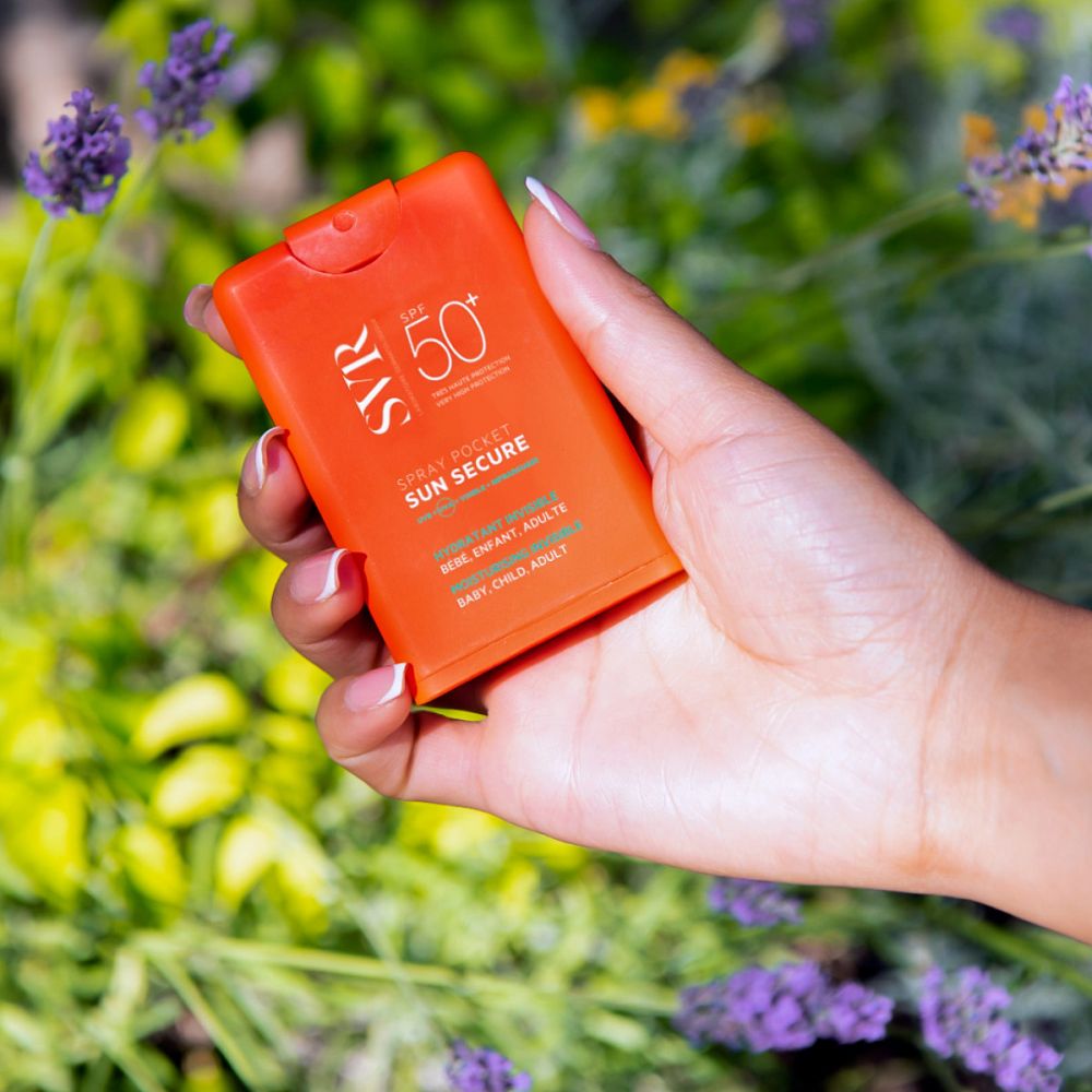 Main tenant un spray de poche SVR SUN SECURE orange. Entouré de fleurs violettes.