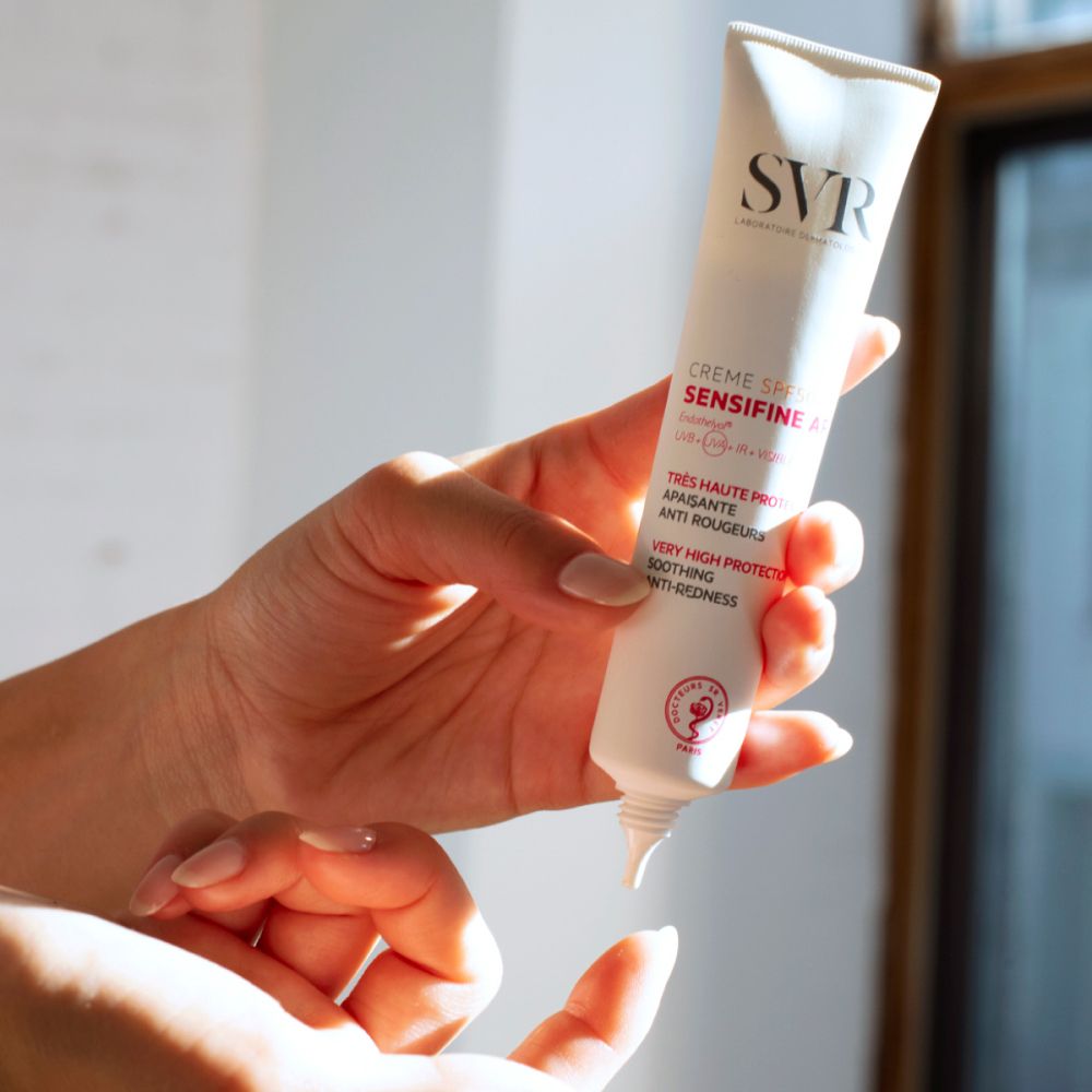 Handen die een SVR Sensifine AR SPF50+ crèmetube vasthouden. Witte tube met rode tekst.