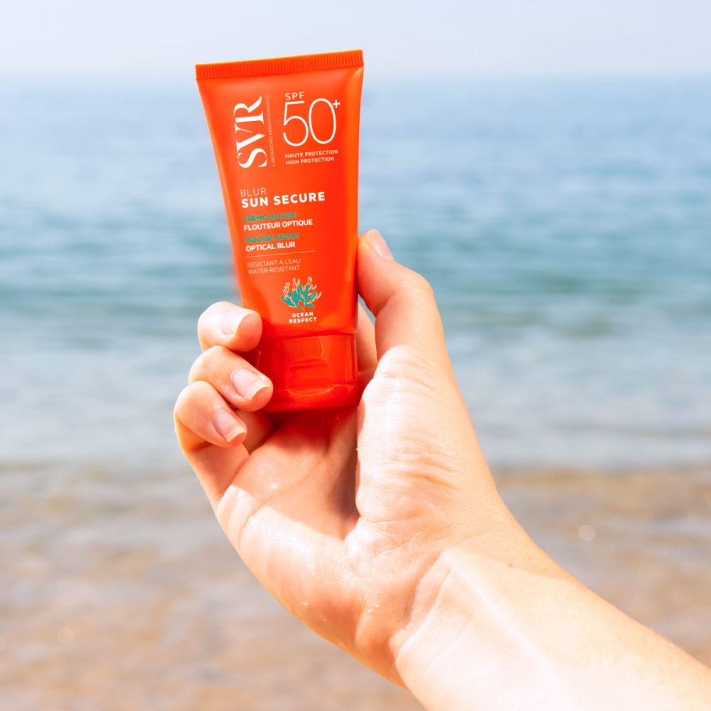 Main tenant un tube orange SVR Sun Secure Blur LSF50+ devant la mer. SPF 50+.
