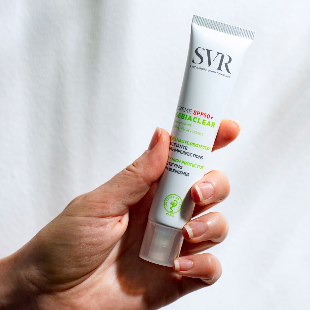 Hand houdt tube vast. SVR-logo. Crème SPF50+ SEBIACLEAR. Groene accenten. Witte tube.