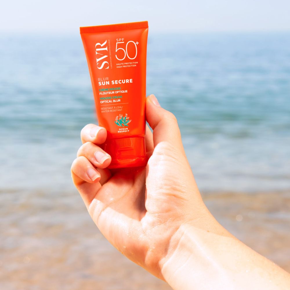 Main tenant un tube orange. Nom du produit: Blur Sun Secure. SPF 50+. Fond: plage et mer.