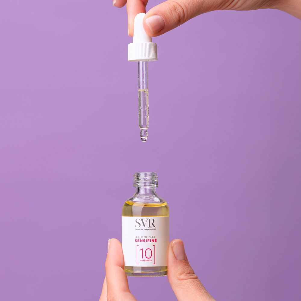 Handen houden een glazen fles met pipet en gele olie. Pipet boven de fles. Opschrift: SVR, Huile de Nuit Sensifine.