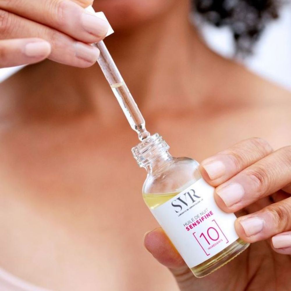 Handen houden een glazen fles met pipet en gele olie. Pipet boven de fles. Opschrift: SVR, Huile de Nuit Sensifine.