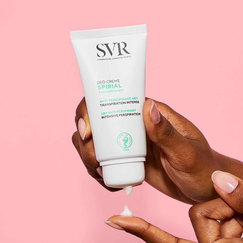 Main tenant un tube SVR SPIRIAL Deo-Creme. Crème appliquée sur le doigt. Fond rose.