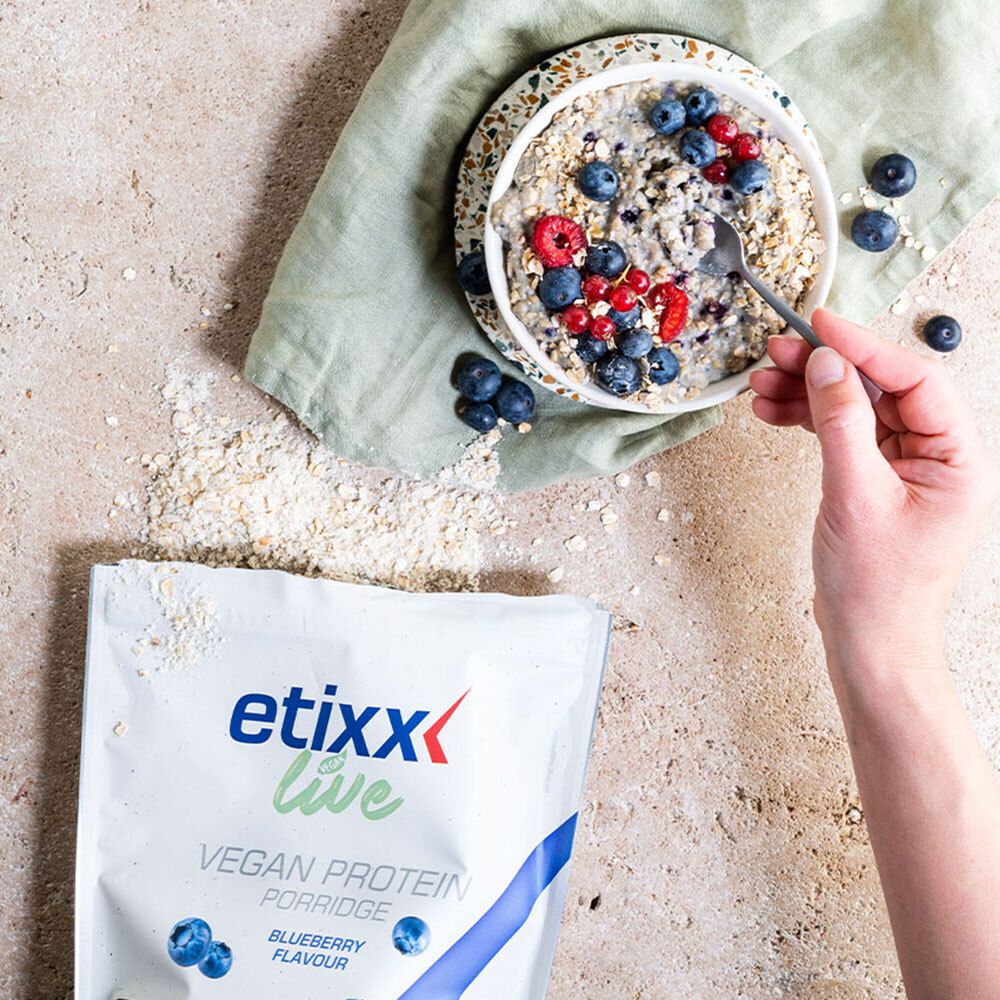 Bol de porridge avec myrtilles et framboises. Main avec cuillère. Sachet 'etixx live Vegan Protein Porridge Blueberry' à côté. Flocons d'avoine dispersés.