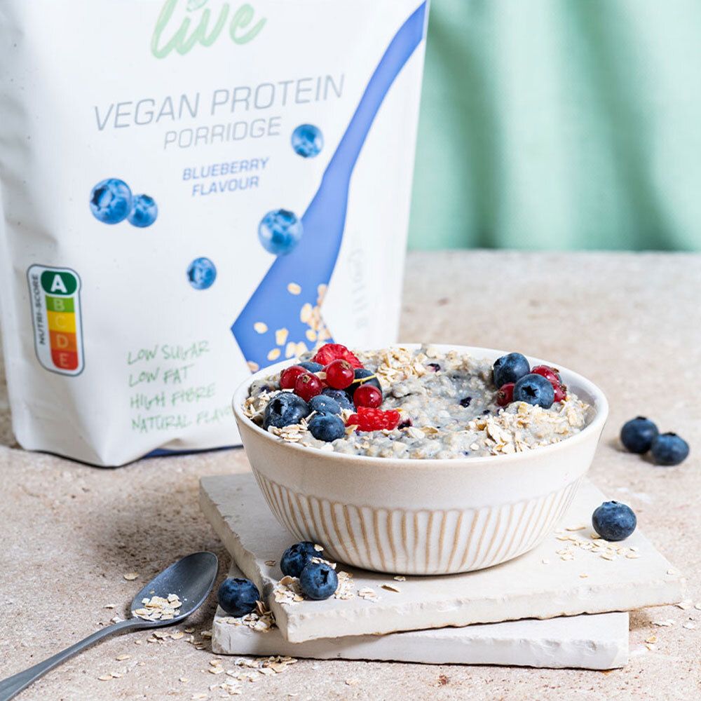 Bol de porridge avec myrtilles et framboises. Sachet 'etixx live Vegan Protein Porridge Blueberry' en arrière-plan. Cuillère et flocons d'avoine à côté.
